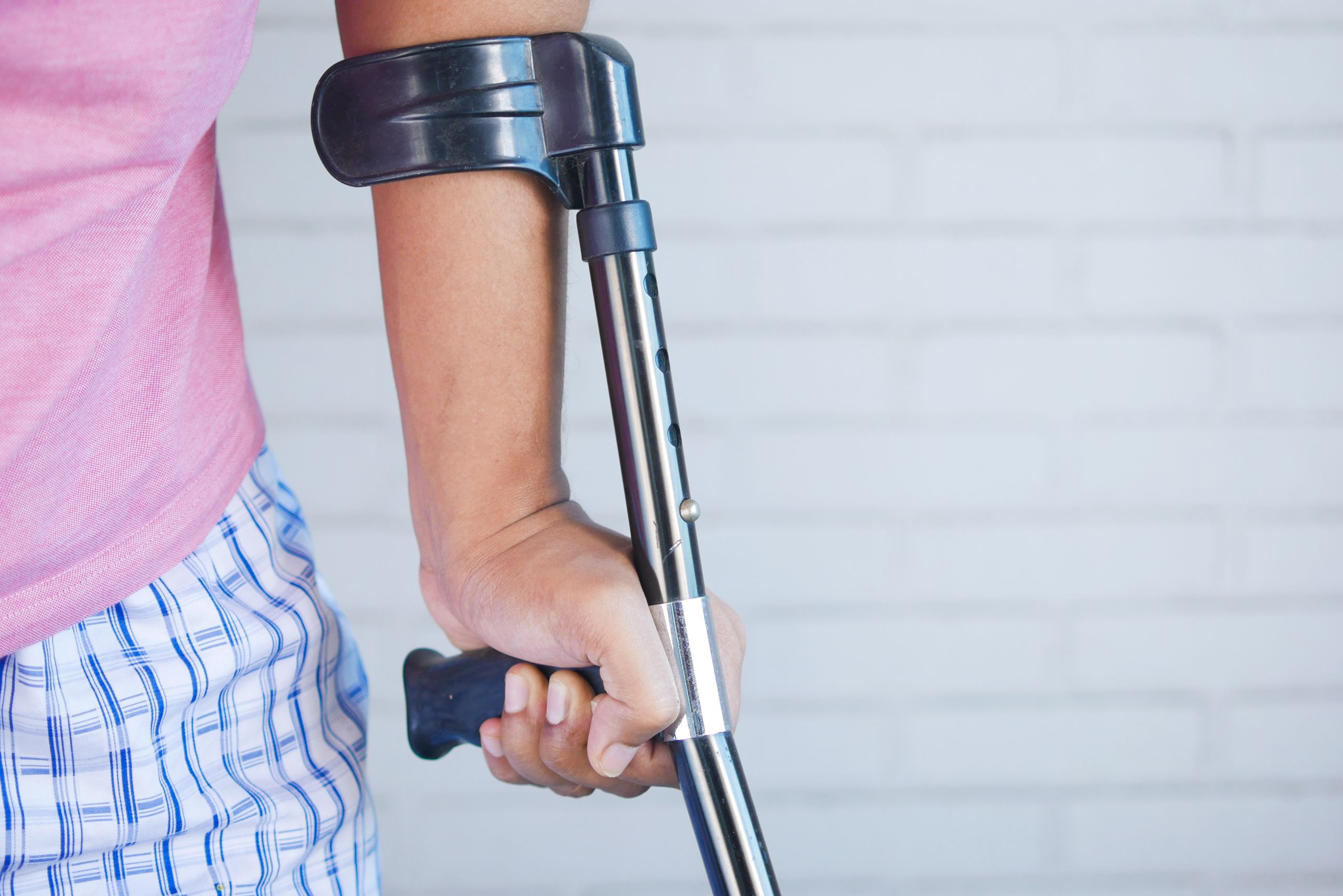 Close-up of a person in a pink shirt using a crutch for support indoors against a brick wall.